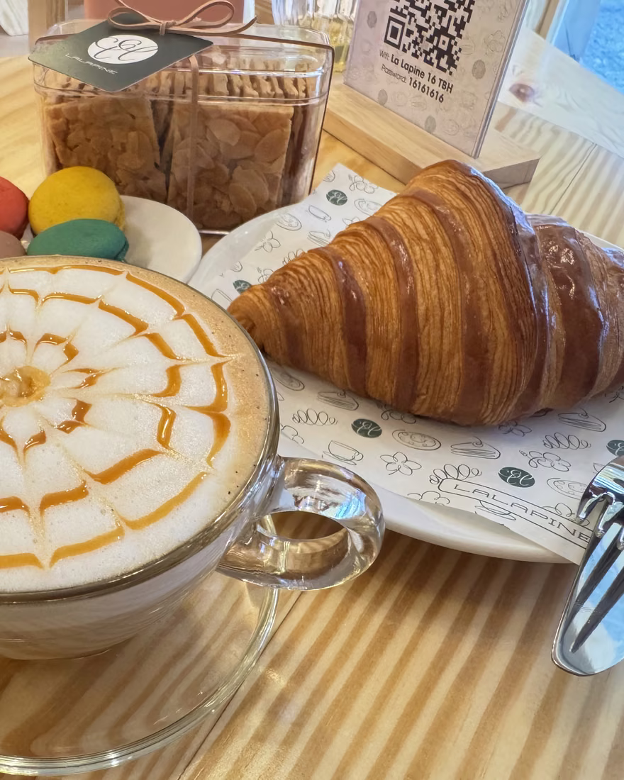 Ly caramel macchiato vẽ hình trên bọt sữa, bên cạnh croissant và ba chiếc macaron