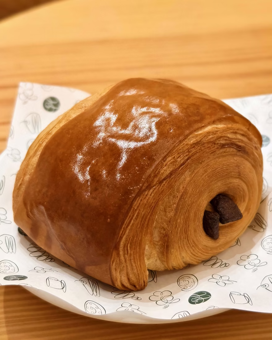 Ba chiếc pain au chocolat trên khay gỗ, kèm biển tên món